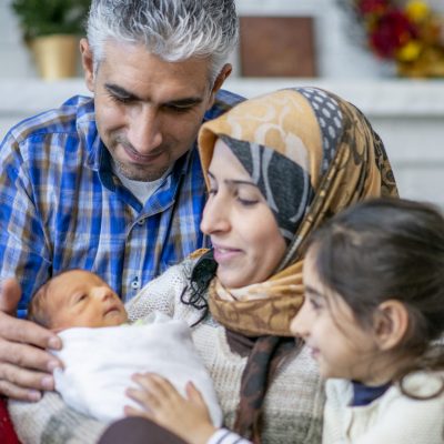 A Muslim family is together in a living room. A mother, father, and daughter are all watching over their newest family member. The newborn baby is sleeping in his mother's arms.